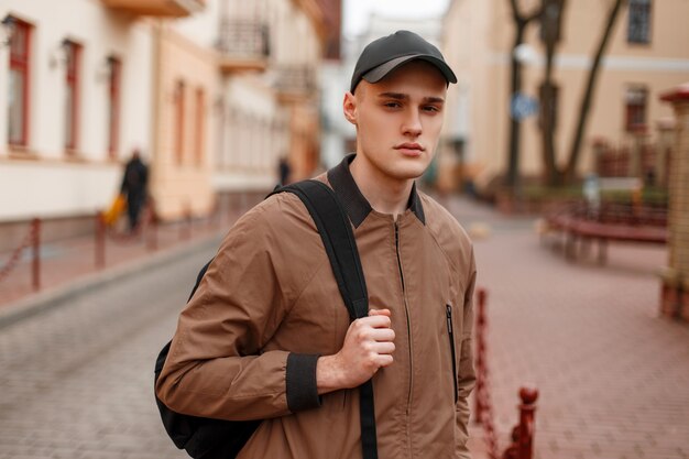 Portrait of a young stylish guy in a fashionable spring beige jacket in a stylish black cap with a sporty trendy backpack in the city