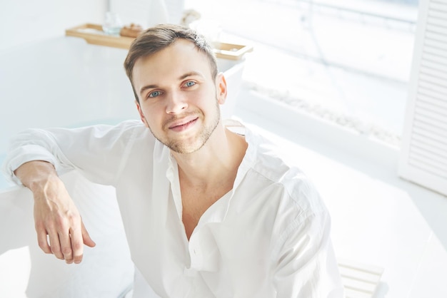 Portrait of a young successful happy man in light clothes sits in his luxury apartment in a white interior and smiles