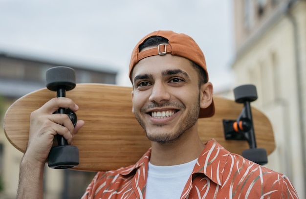 Portrait of young successful skater holding longboard, walking on the street, smiling