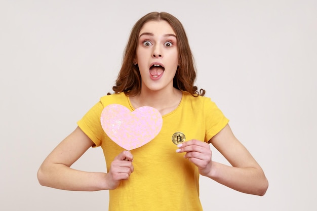 Портрет молодой удивленной женщины в желтой повседневной футболке с розовым сердцем на палочке и физическим биткойном шокировал выражение лица криптовалюты Крытый студийный снимок изолирован на сером фоне