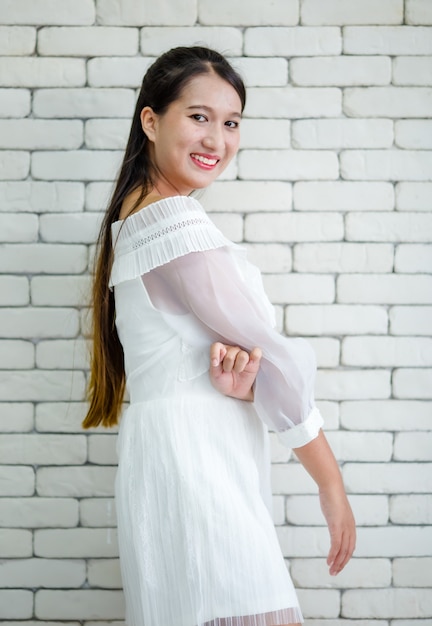 Portrait of young teen Asian female in white dress standing on background of white brick wall with self confident and positive smiling pose.