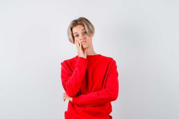 Portrait of young teen boy leaning cheek on hand in red sweater and looking pensive front view