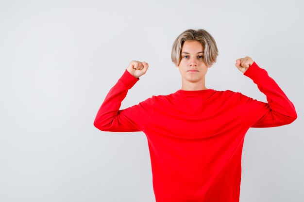 Портрет молодого мальчика-подростка, показывающего мышцы рук в красном свитере и уверенно выглядящего, вид спереди