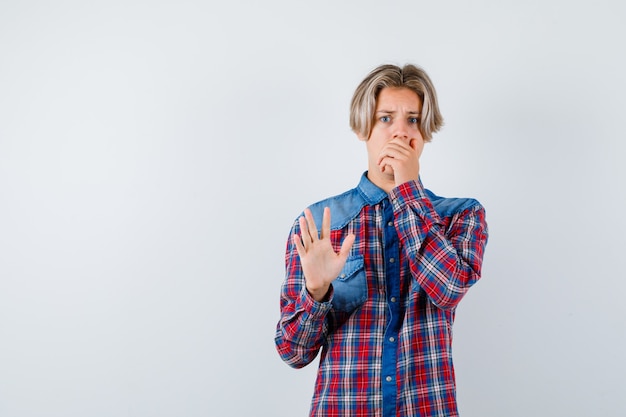 Portrait of young teen boy showing stop gesture, keeping hand on mouth in checked shirt and looking scared front view