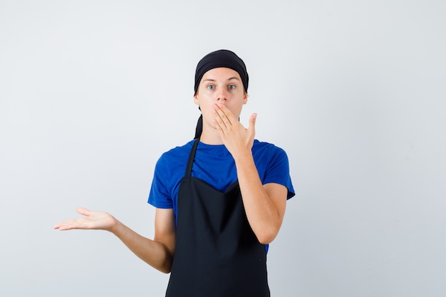 Portrait of young teen cook with hand on mouth, spreading palm aside in t-shirt, apron and looking surprised front view