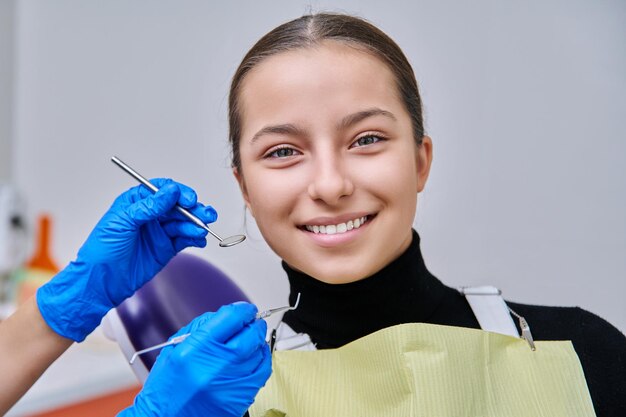 Portrait of young teenage girl in dental chair with hands of doctor with tools Female teenager smiling with teeth looking at camera in dentist office Adolescence hygiene treatment dental health care