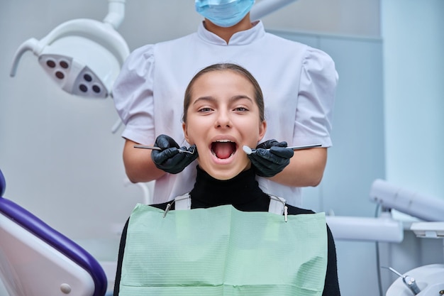 ツールを持つ医師の手で歯科椅子に座っている若い 10 代の少女の肖像画歯科医院でカメラを見て歯で微笑む女性 10 代の少女青少年の衛生治療歯科医療