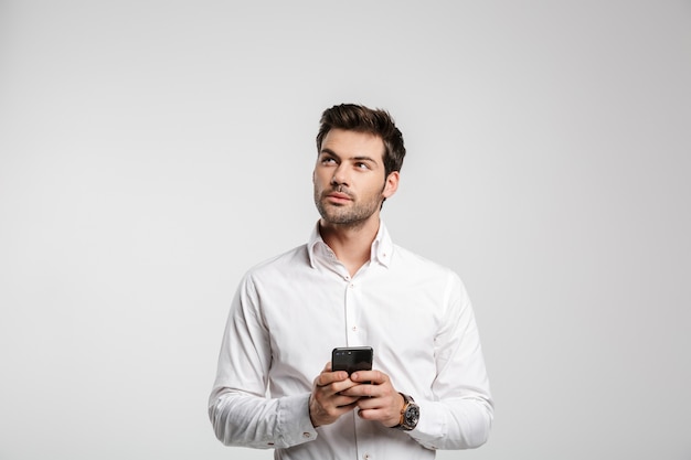 Portrait of young thinking businessman in wristwatch typing on cellphone and looking upward isolated on white