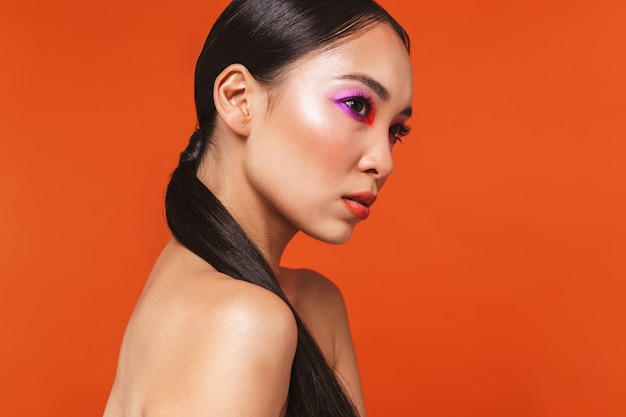 portrait of a young topless asian woman with brunette hair wearing bright makeup, standing isolated on red, looking away, posing