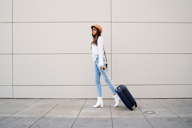 Portrait of young traveler woman carrying a suitcase while walking outdoors on the street. Tourism concept.