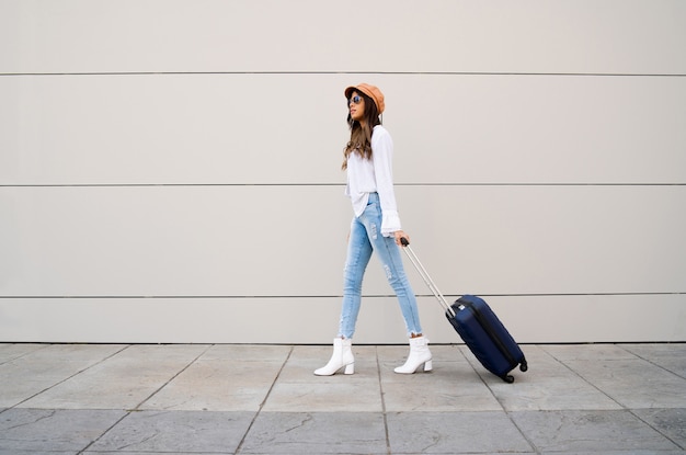 Portrait of young traveler woman carrying a suitcase while walking outdoors on the street