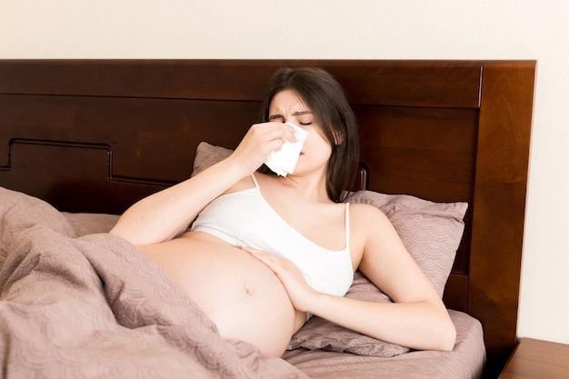 Portrait of young unhappy pregnant woman leaning on pillows on bed and blowing her nose into tissue Future mom caught cold and sneezing
