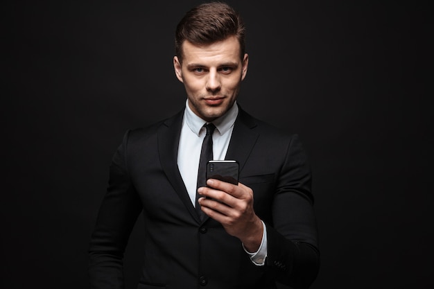 Portrait of young unshaven businessman dressed in formal suit using cellphone and looking at camera isolated over black wall