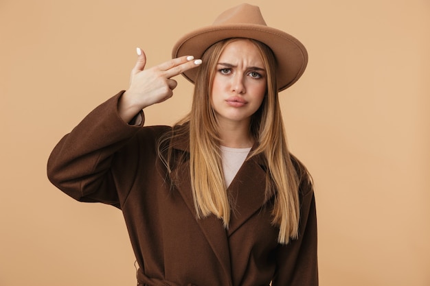 Portrait of young uptight girl wearing hat and coat holding gun fingers at her temple as killing herself isolated on beige