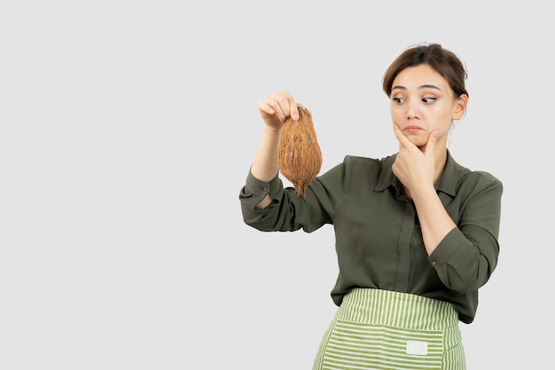 Portrait of young woman in apron holding a coconut against white wall