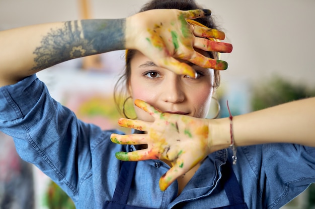 Portrait of young woman in apron looking at camera and showing her hands in paint while painting