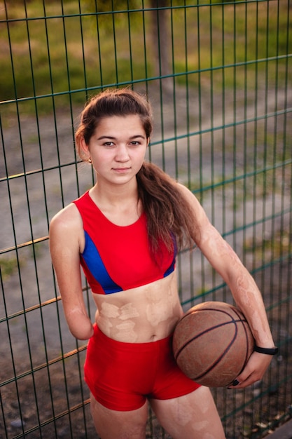 Foto atleta della giovane donna del ritratto con un braccio amputato e ustioni sul suo corpo tiene nelle mani la palla da basket dopo l'allenamento all'aperto al tramonto