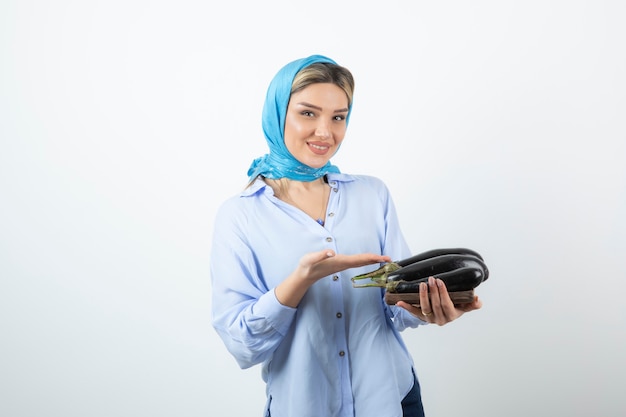 Portrait of young woman in blue shawl holding wooden board of uncooked eggplants