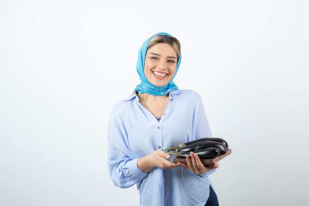 Portrait of young woman in blue shawl holding wooden board of uncooked eggplants