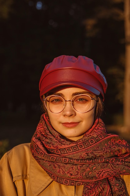 Portrait of young woman in cap and scarf standing in autumn park at sunset and looking at camera