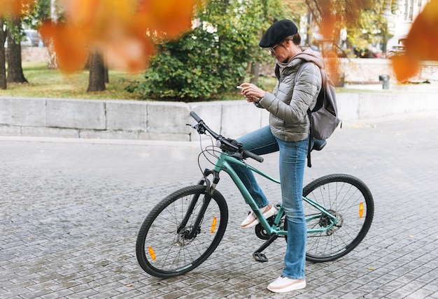 Foto ritratto di una giovane donna con il berretto e gli occhiali da sole che usa il cellulare in bicicletta in una soleggiata giornata d'autunno in città