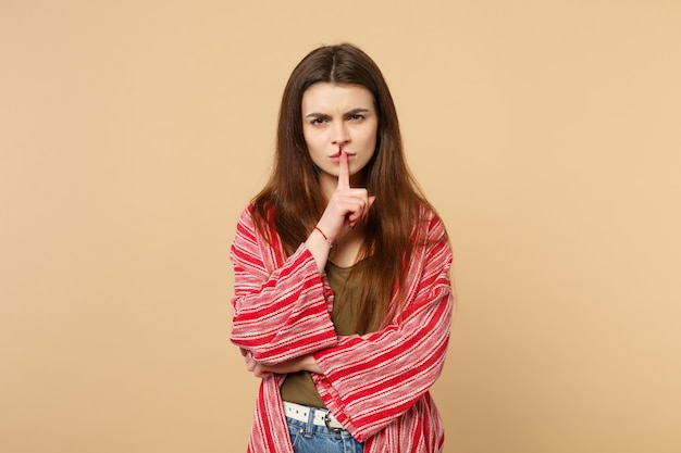 Portrait of young woman in casual clothes saying hush be quiet with finger on lips shhh gesture isolated on pastel beige wall background. People sincere emotions lifestyle concept. Mock up copy space.