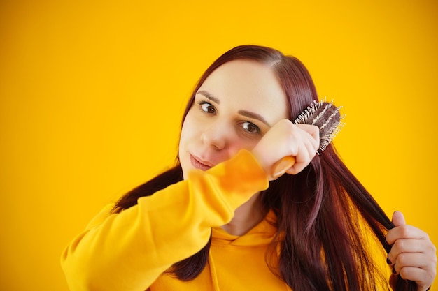 Portrait of young woman combing her hair on yellow background Brunette in yellow hoodie preens while looking at camera