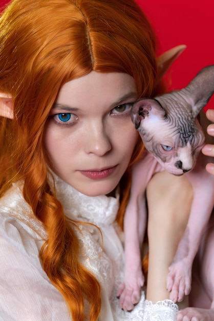 Photo portrait of young woman cosplay elf with red hair in white dress holding half asleep sphinx kitten