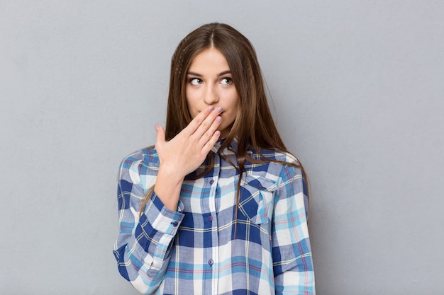 Portrait of a young woman covering her mouth with palm and looking away over gray wall