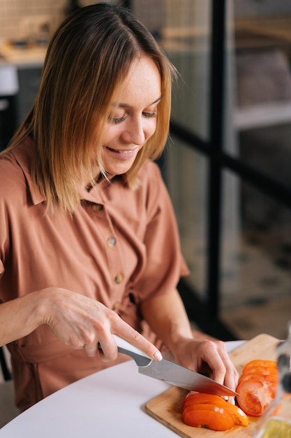 Portrait of young woman cutting fresh tomato using kitchen knife on wooden cutting board. Young woman cutting fresh organic tomato with a knife for vegetable salad. Concept of healthy eating.