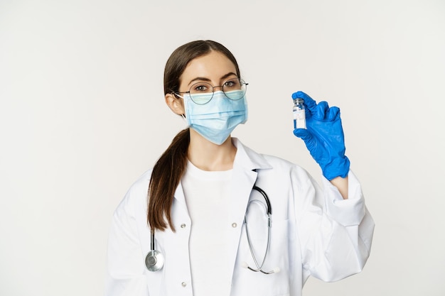 Portrait of young woman doctor in medical face mask and uniform showing vaccine covid vaccination ca...