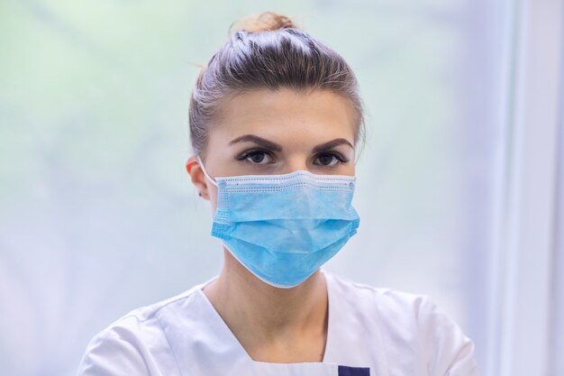 Portrait of young woman doctor nurse in protective medical face mask, background interior of clinic