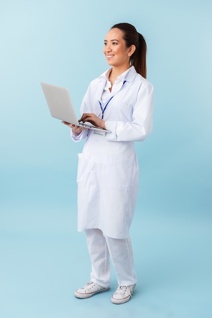 ラップトップコンピューターを使用して青い壁の上に孤立してポーズをとる若い女性医師の肖像画。