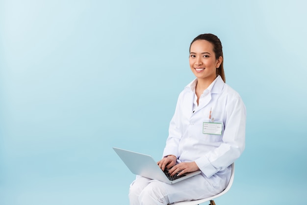 portrait of a young woman doctor posing isolated over blue wall using laptop computer.