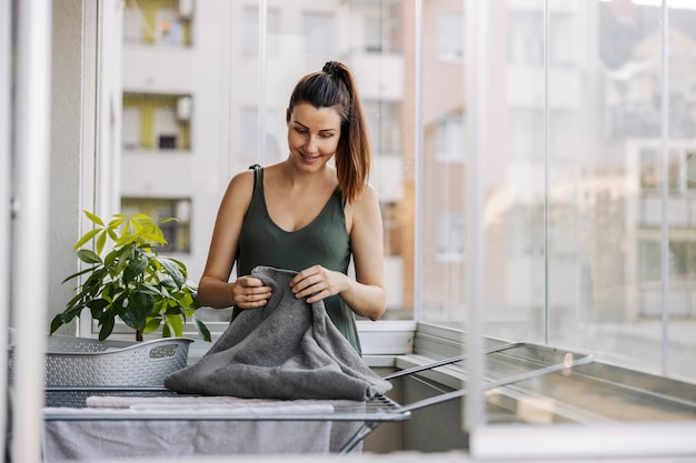 Portrait of a young woman doing chores, stacking and spreading laundry on the balcony. A smiling woman dressed in casual clothes puts towels on the terrace on a sunny summer day