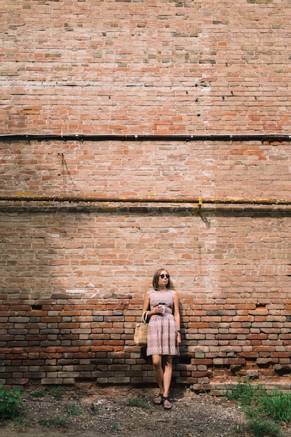 Portrait of young woman in dress and sunglasses leaning on brickwall with coffee cup