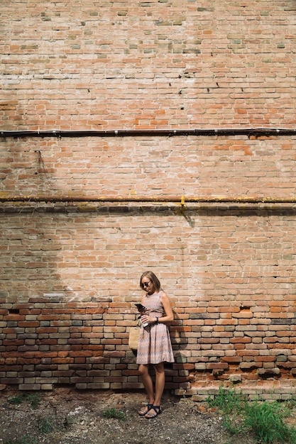 Portrait of young woman in dress and sunglasses leaning on brickwall with coffee cup