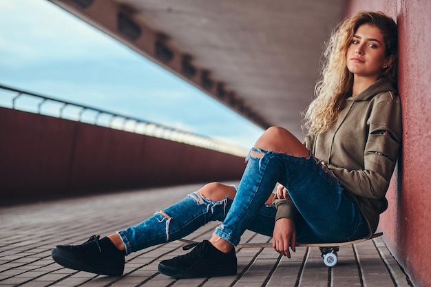 Portrait of a young woman dressed in a hoodie and ripped jeans leaning on a wall while sitting on a skateboard at bridge footway.