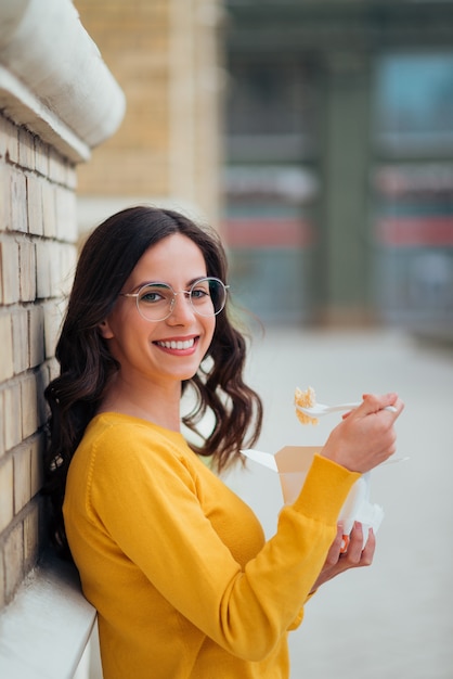 Portrait of a young woman eating from takeaway box outdoors, leaning on a wall and smiling at camera.