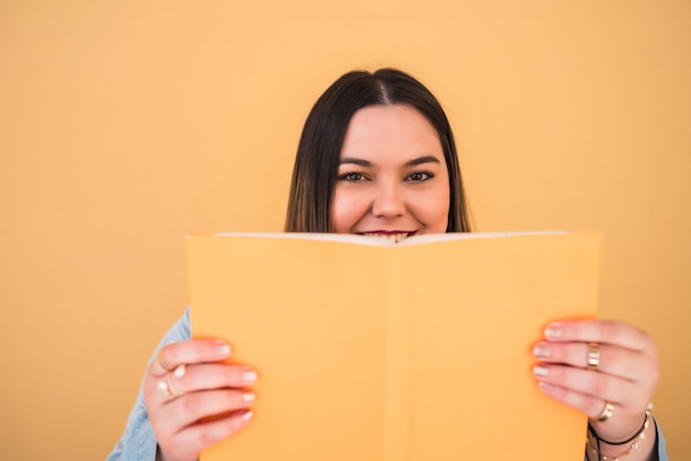 Portrait of young woman enjoying free time and reading a book while standing against yellow wall