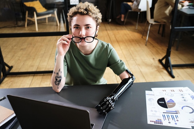 Portrait of young woman in eyeglasses with prosthetic arm looking at camera while working on laptop at desk