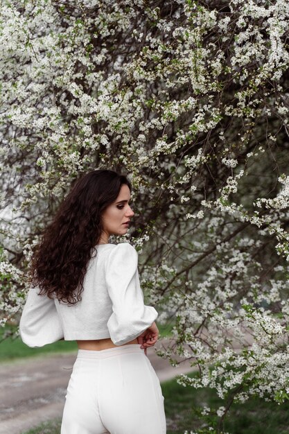 Portrait of young woman in the garden. Attractive girl weared white dress posing near blooming trees.