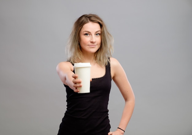 Portrait of young woman giving hot coffee or tea beverage to camera against gray background