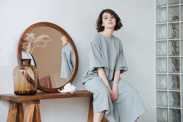 Portrait of a young woman in a grey dress sitting on a table with a round mirror. Her profile reflecting in the mirror. Beautiful brunette with short hair indoors.