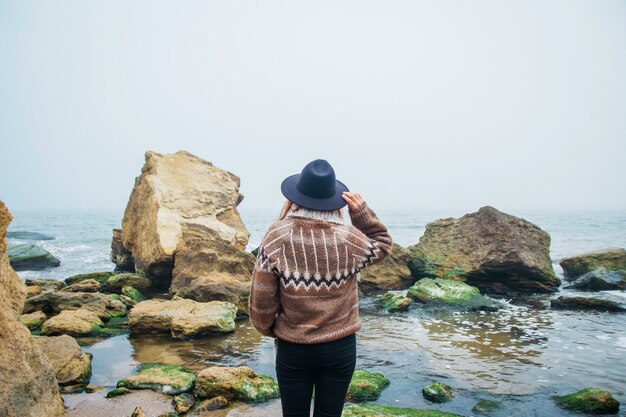 美しい海を背景に岩の上に帽子をかぶった若い女性の肖像画後ろから撮影