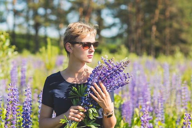 Portrait of young woman holding bouquet of lupin flowers walking in summer meadow Stylish girl is enjoying sunny weather blonde woman with bouquet of flowersLupine field Summer vacation