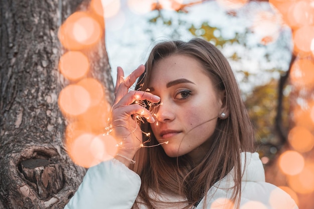Portrait of young woman holding illuminated string light by tree