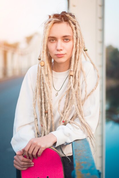 Foto ritratto di una giovane donna che tiene uno skateboard mentre si trova su un ponte pedonale durante il tramonto