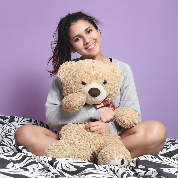 Portrait of young woman holding toy while sitting on bed against wall