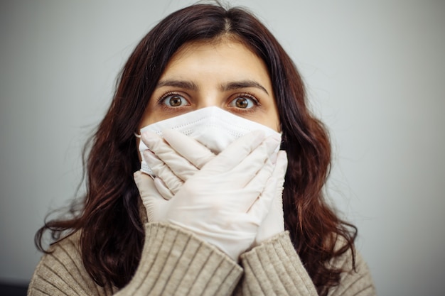 Photo portrait of young woman holds hands on her mouth showing silence sign and stays home during quarantine due to coronavirus pandemia. beautiful girl wearing medical mask and gloves. covid-19 epidemia.